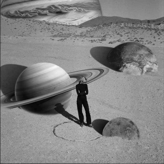 A surreal black-and-white digital collage shows a man standing in a desert landscape alongside giant planets resembling Saturn and Jupiter, with his shadow forming a long streak across the sand.