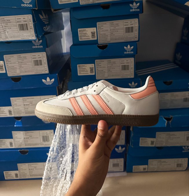 A hand holding up a white Adidas Samba OG sneaker with pink/coral colored stripes and heel tab, displayed in front of stacked blue Adidas Originals shoe boxes.