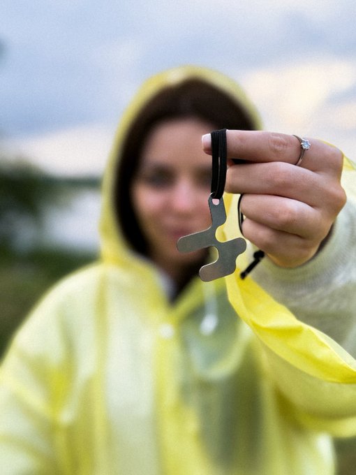 A photograph of a smiling person wearing a bright yellow hooded raincoat, holding up a small silver metal tool attached to a black lanyard.