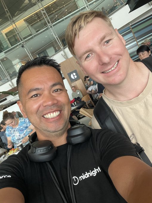 A selfie taken in an airport or terminal showing two smiling men, one wearing a black 'midnight' t-shirt with Sony headphones around his neck, and the other in a tan t-shirt.