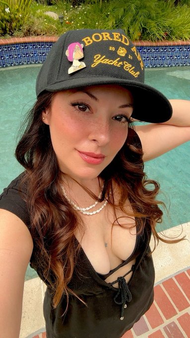 A selfie of a woman wearing a black 'Bored Yacht Club' cap with a pink skull design, a black top, and pearl necklace, posing by a swimming pool.