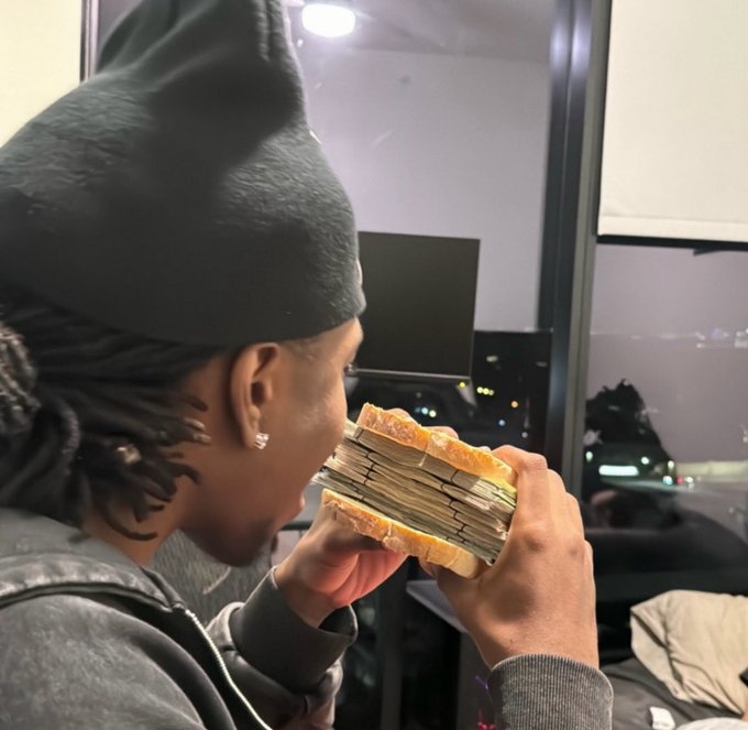 A person in a black beanie biting into a thick stack of cash held in their hands, with a city view visible through a window in the background.