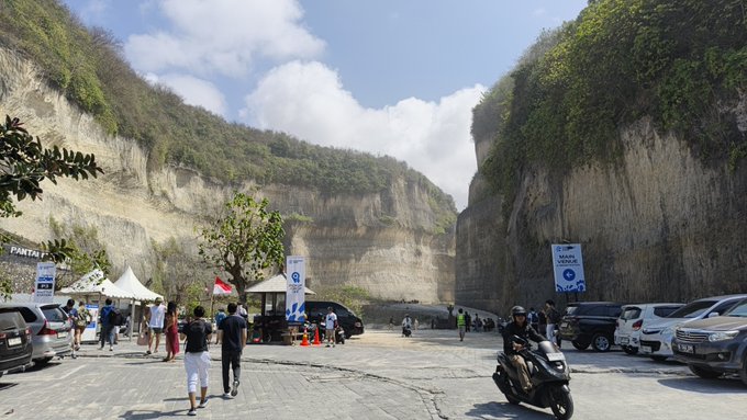 A bustling outdoor scene at a quarry cliff location with people walking and a motorcyclist, featuring tents, parked cars, and a 'PANTAI' sign on the left.