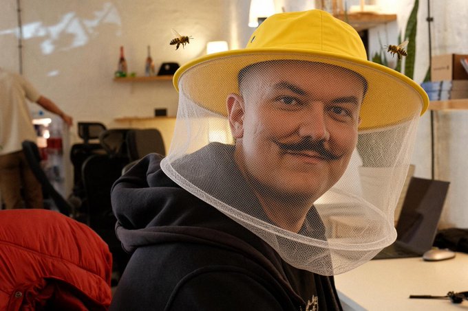 A man with a mustache smiles at the camera while wearing a yellow beekeeper's hat with a mesh veil, with two bees visible flying near the mesh.
