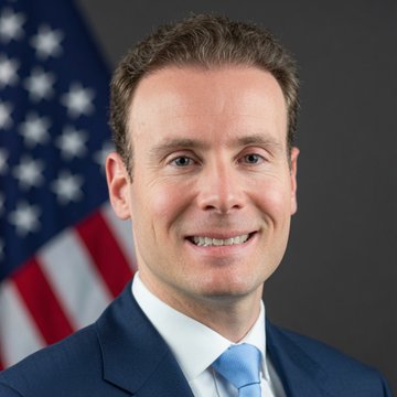 An official portrait of a man in a dark suit and blue tie smiling, with an American flag visible in the background.