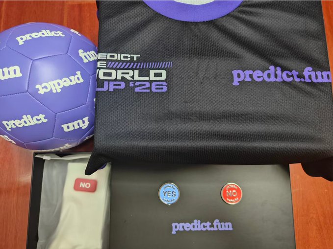 Photograph of Predict.fun promotional merchandise including a purple soccer ball, a 'WORLD CUP '26' t-shirt, and YES/NO pin buttons in a box.