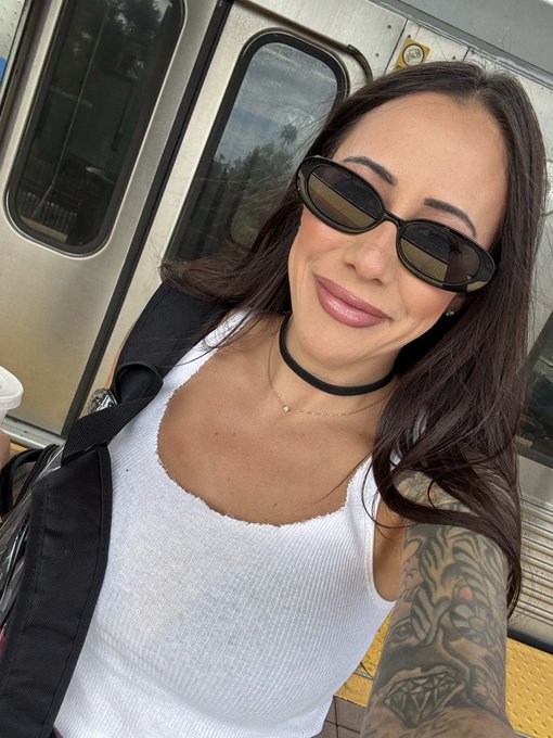 A young woman with dark hair, oval sunglasses, a black choker, a white tank top, and a colorful sleeve tattoo takes a selfie on what appears to be a Metro train platform, captioning it "I'm taking public transportation in LA on a Saturday night and that is my thesis on why all coins go HIGHER" — humorously implying that even crypto traders are out clubbing and not trading, so the market is sure to pump.