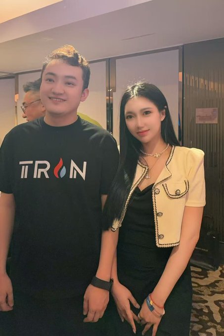 Justin Sun, wearing a black 'TRON' t-shirt, poses for a photo with a young woman wearing a black dress and a cream-colored jacket with black trim.