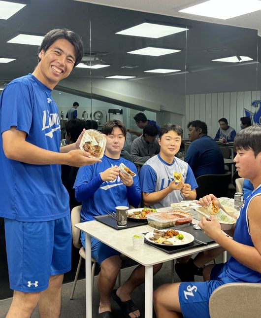 Members of what appears to be a baseball team in blue uniforms celebrate their first victory together by enjoying a meal, with one player (likely Satoshi) sharing tacos with teammates in a team dining area. The players are smiling and holding food items like tacos and skewers, reflecting the post's caption about celebrating their win with 80 tacos shared among grateful teammates.