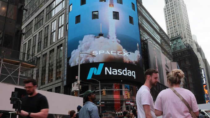 A large digital billboard in Times Square displays a SpaceX rocket launch with the "Nasdaq" logo, while pedestrians walk by on the street below.