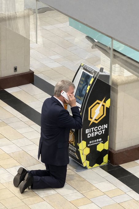 The image depicts a man kneeling down while talking on his cell phone, surrounded by white tiles, with an advertisement for Bitcoin Depot prominently displayed above him. The text on the sign indicates that the coin is being sold at the average price of $28,815. This suggests that the man could be interested in purchasing Bitcoins or checking the market conditions related to Bitcoin transactions.