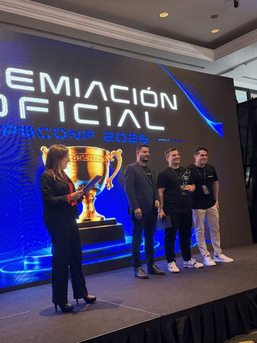 Four people stand on a stage at a conference in front of a large screen displaying 'PREMIACIÓN OFICIAL' and '...DBCONF 2026' alongside an image of a golden trophy.