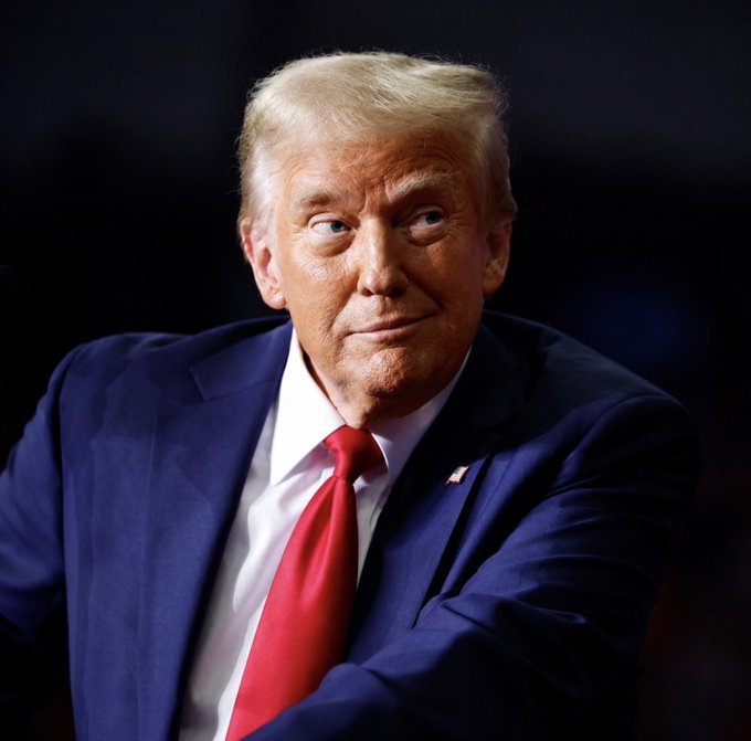 A portrait photograph of Donald Trump wearing a navy blue suit with a red tie and an American flag pin, looking off to the side.