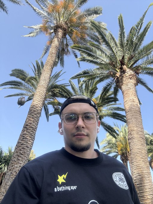 A young man wearing a black cap, sunglasses, and a black 'a bathing ape' t-shirt takes a low-angle selfie beneath tall palm trees against a clear blue sky.