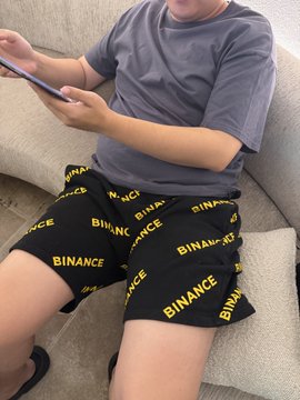 A young person sitting on a couch holding a smartphone, wearing a grey t-shirt and black shorts branded with the yellow 'BINANCE' logo.
