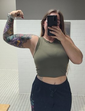 A woman takes a mirror selfie in a tiled bathroom, wearing an olive green crop top and black sweatpants, showing off her colorful full-sleeve tattoo and another tattoo on her waist.
