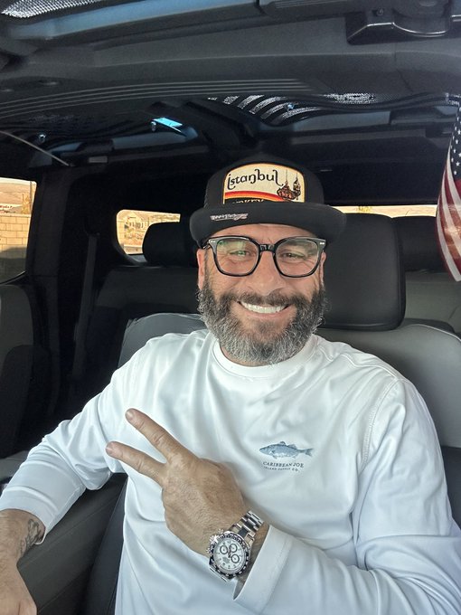 A smiling, bearded man wearing glasses and a 'Starbu' cap poses inside a vehicle, wearing a white long-sleeve shirt featuring a 'Carpenter Joe' logo and a silver watch.