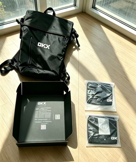 An unboxed black OKX backpack with its packaging and accessories arranged on a wooden floor.