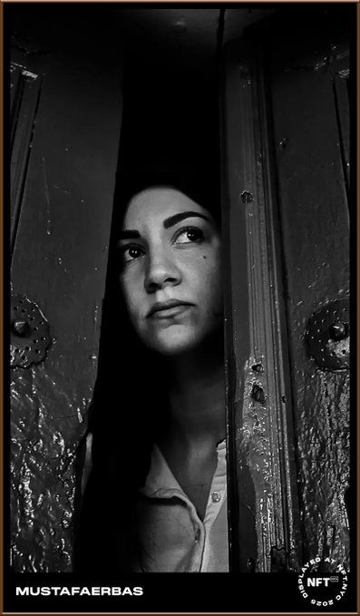 A black and white portrait of a woman peering through a partially open door, with 'MUSTAFAERBAS' watermark and an 'NFT' logo in the bottom corners.