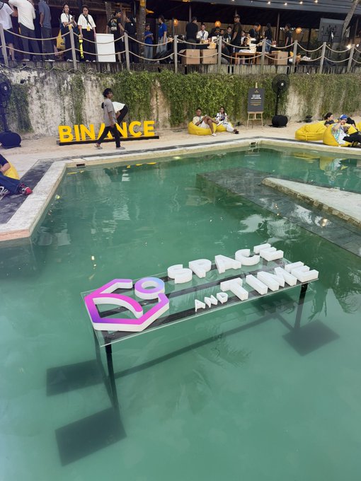 An outdoor pool venue with a 'BINANCE' sign in yellow letters and a '69 SPACE AND TIME' sign standing in the water, with people gathered around the pool area.