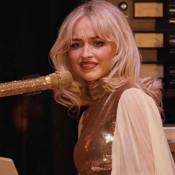 A blonde woman with wavy hair, wearing a gold sequin top, sits in front of a gold microphone with a displeased expression.