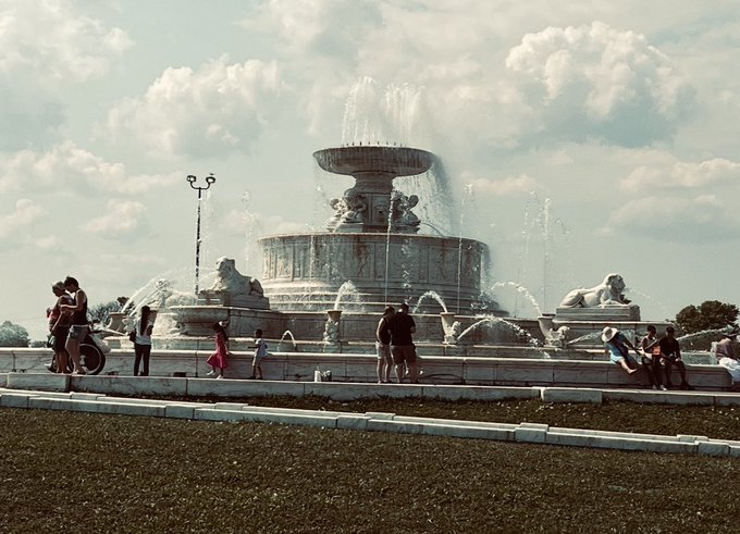 A wide shot of a large, ornate multi-tiered stone fountain in a public plaza under a partly cloudy sky, with several people gathered around its base.