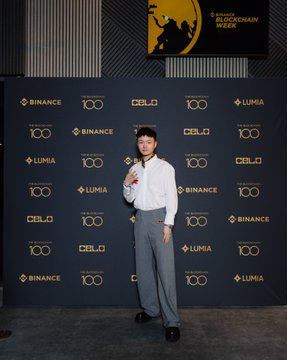 A man in a white shirt and gray trousers poses at a press backdrop featuring "BINANCE," "CELO," "LUMIA," and "Binance Blockchain Week" logos, which ties into the caption's mention of "big sis @heyibinance" hosting a formal awards ceremony at the glamorous Binance Blockchain Week event.