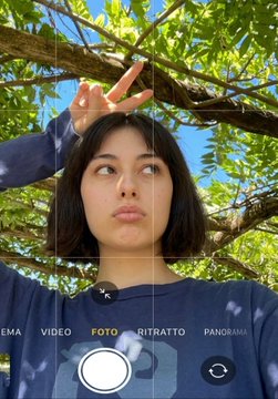 A selfie of a young woman outdoors under a tree, making a peace sign, displayed within a phone camera interface showing modes like 'VIDEO', 'FOTO', and 'RITRATTO'.
