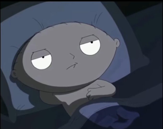 Stewie Griffin from Family Guy lies in bed with a half-asleep, unamused expression under a blue blanket.