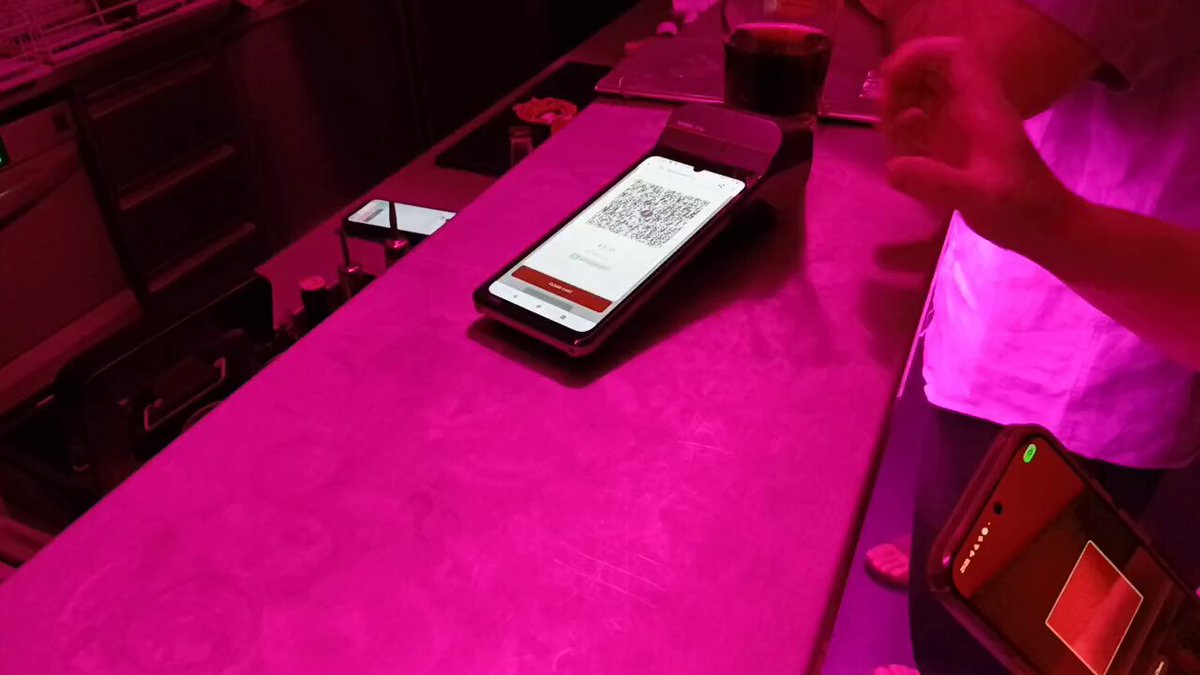 The image shows a person holding a smartphone with a QR code displayed on its screen. The QR code appears to be related to Ark #bitcoin payments or a similar payment system. There are two other people present, one of whom is using a credit card machine at a bar counter. A cup can also be seen in the scene.