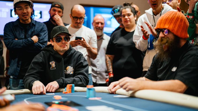 Two poker players sit across from each other at a blue felt table, surrounded by spectators, with one player wearing a black hoodie and cap and the other sporting an orange beanie and a thick beard.