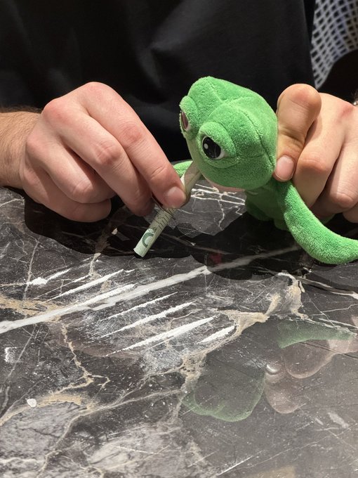 A pair of hands uses a green marker to draw on a black marble countertop next to a small green lizard plush toy.