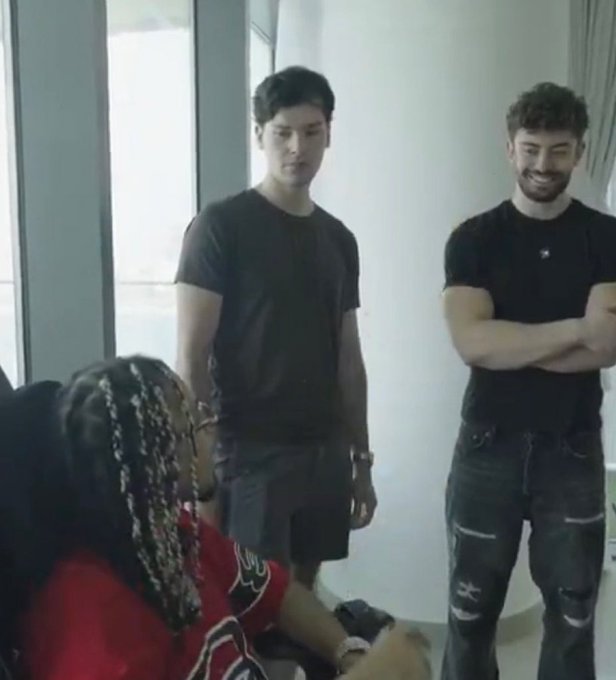 Two men stand smiling in a modern room—one in a plain gray t-shirt, the other in a black shirt and ripped jeans—while a seated person with braided hair in a red shirt looks up at them. The relaxed, celebratory vibe of the group appears to reflect the milestone mentioned in the post of hitting a $200K market cap.