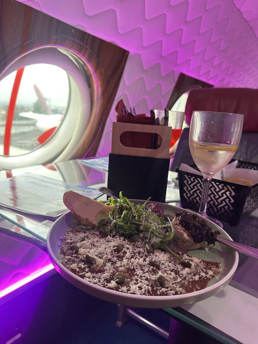 A meal is served on a plane with a distinctive window and pink ambient lighting, featuring a pasta dish with greens and grated cheese alongside a glass of white wine.