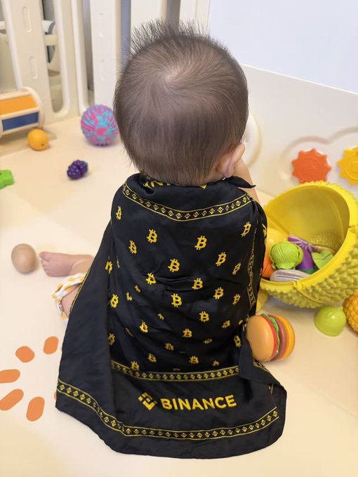 A baby sits on a play mat surrounded by colorful toys, draped in a black cape featuring the Bitcoin "₿" logo and the word "BINANCE" in yellow. The image playfully ties into the post's theme of early crypto investment, humorously suggesting the child is being prepared as a future "old driver" of the Binance and crypto markets.