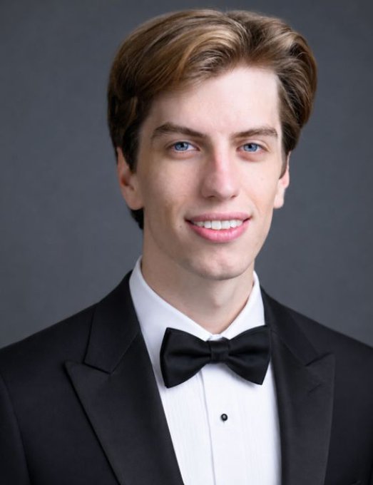 A formal portrait photo of a young man with light brown hair and blue eyes, wearing a black tuxedo with a white shirt and black bow tie against a gray background.