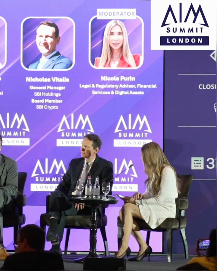 The image shows a group of people sitting around a table with chairs, engaged in conversation. There are two men and one woman present in the scene. The man on the left side of the image appears to be wearing a suit and tie, while the other man is dressed more casually. The woman sits next to them at the table.

The background features a purple wall adorned with various logos, including "AIM" and "blockchain". There are also several bottles placed around the room, possibly as refreshments for the gathering.