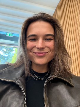 A young woman with long, tousled brown hair smiles warmly at the camera in a selfie taken indoors, wearing a black leather jacket and layered silver necklaces, likely at the Bitpanda office in Vienna.