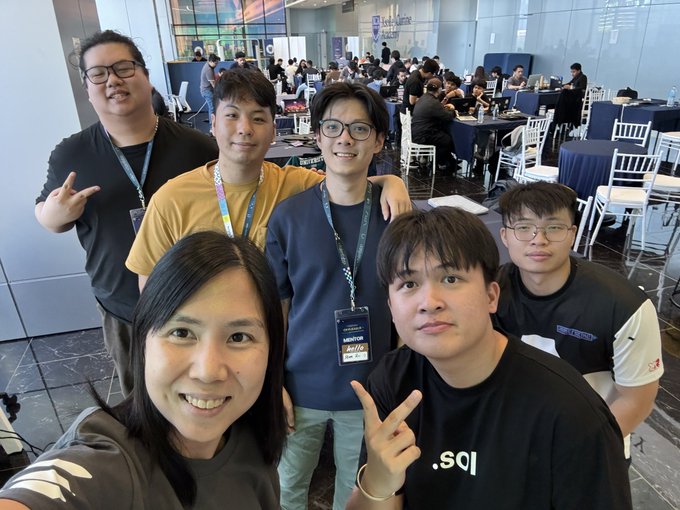 A group selfie of six smiling people posing together in a conference hall with tables and chairs visible in the background.