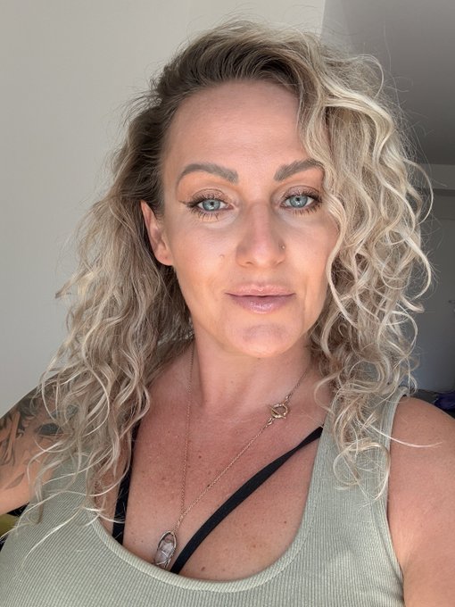 A close-up selfie shows a woman with curly blonde hair, blue eyes, and a nose piercing, wearing a light green tank top and a pendant necklace.