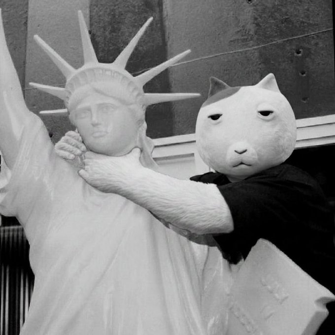 A black-and-white photo of the Cat Meme (CatBenatar) hugging a replica of the Statue of Liberty in an awkward embrace.