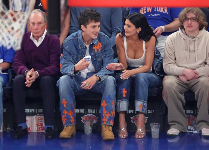 Timothée Chalamet sits courtside at a New York Knicks game wearing a denim outfit with orange cross designs, sitting next to Kylie Jenner in a denim and white outfit.