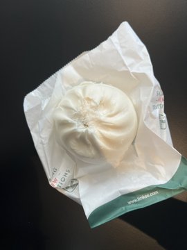 A white steamed bun (likely a baozi) wrapped in paper from a shop called 'Bun Bun' on a dark surface.