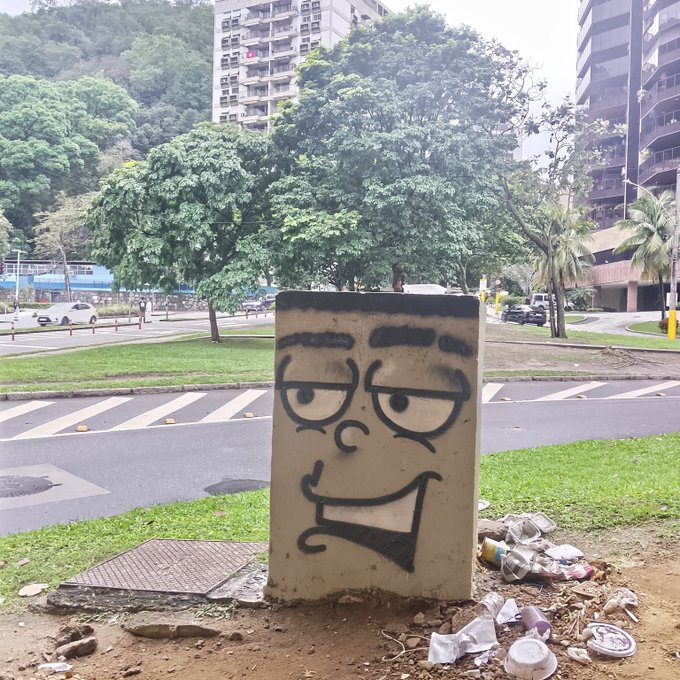 A concrete utility box with a black spray-painted cartoon face (eyes, eyebrows, nose, and mouth) stands on a trash-strewn patch of dirt beside a road in what appears to be Rio de Janeiro, with high-rise buildings and forested hills in the background.