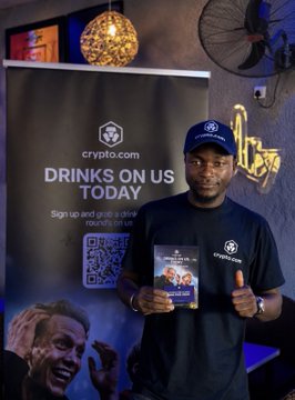 Man wearing a crypto.com branded cap and t-shirt holding promotional flyers in front of a 'crypto.com DRINKS ON US TODAY' banner with a QR code.