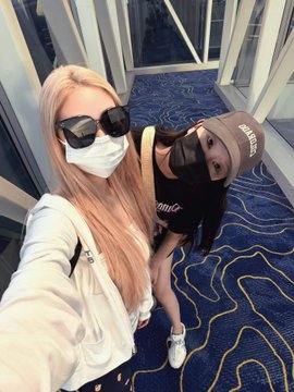 A selfie of two young women wearing face masks and sunglasses, one in a white hoodie and the other in a black shirt and beige cap, standing in a hallway with patterned carpet.
