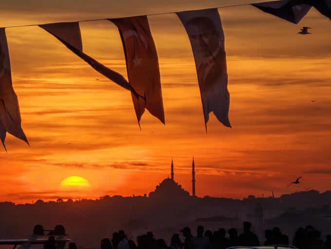 A sunset scene over a city skyline featuring a silhouetted mosque with minarets, framed by Turkish flags and a crowd of people.