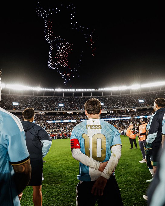 Soccer player Lionel Messi, wearing the number 10 Argentina jersey with his name on the back, stands on the field with his arms behind his back, watching a drone show in the night sky above the stadium.