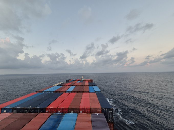 An aerial view from the bridge of a cargo ship loaded with red, blue, and brown shipping containers, sailing on a vast ocean under a cloudy sky.