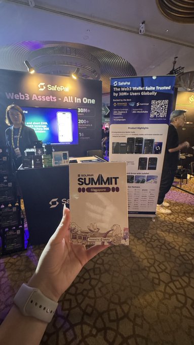 A hand holds a 'SUMMIT Singapore' badge in front of a SafePal booth at a crypto/Web3 conference, displaying banners for 'Web3 Assets - All In One' and 'The Web3 Wallet Suite Trusted by 30M+ Users Globally'.